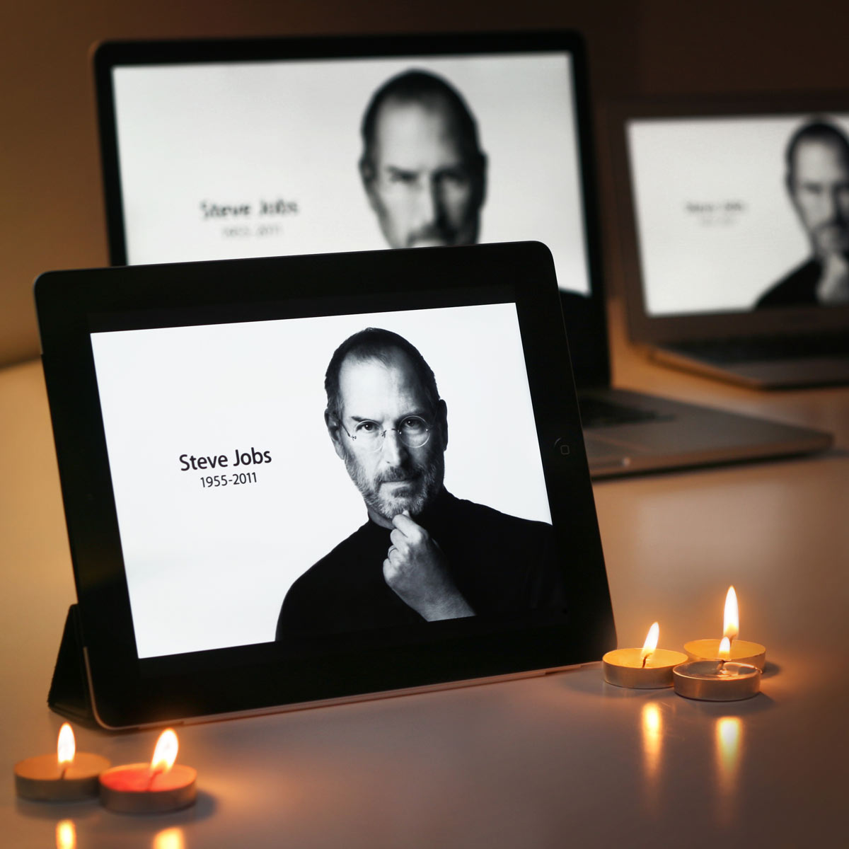 An iPad and two computers with candles in front of them with Steve Jobs on the screens